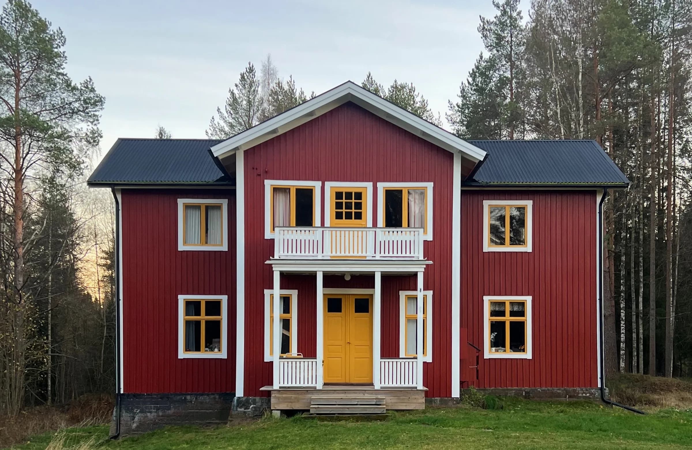 Rött trähus med gula fönsterbågar, vit veranda och balkong omgivet av skog